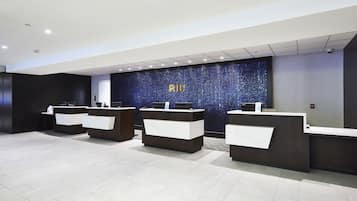 Een receptiehal met een donkerblauwe wand en de letters 'RIU' in goud. Er zijn drie receptiebalies met witte bladen en donkere onderstellen.