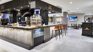 A modern hotel bar with a counter displaying pastries and a digital screen in the background.