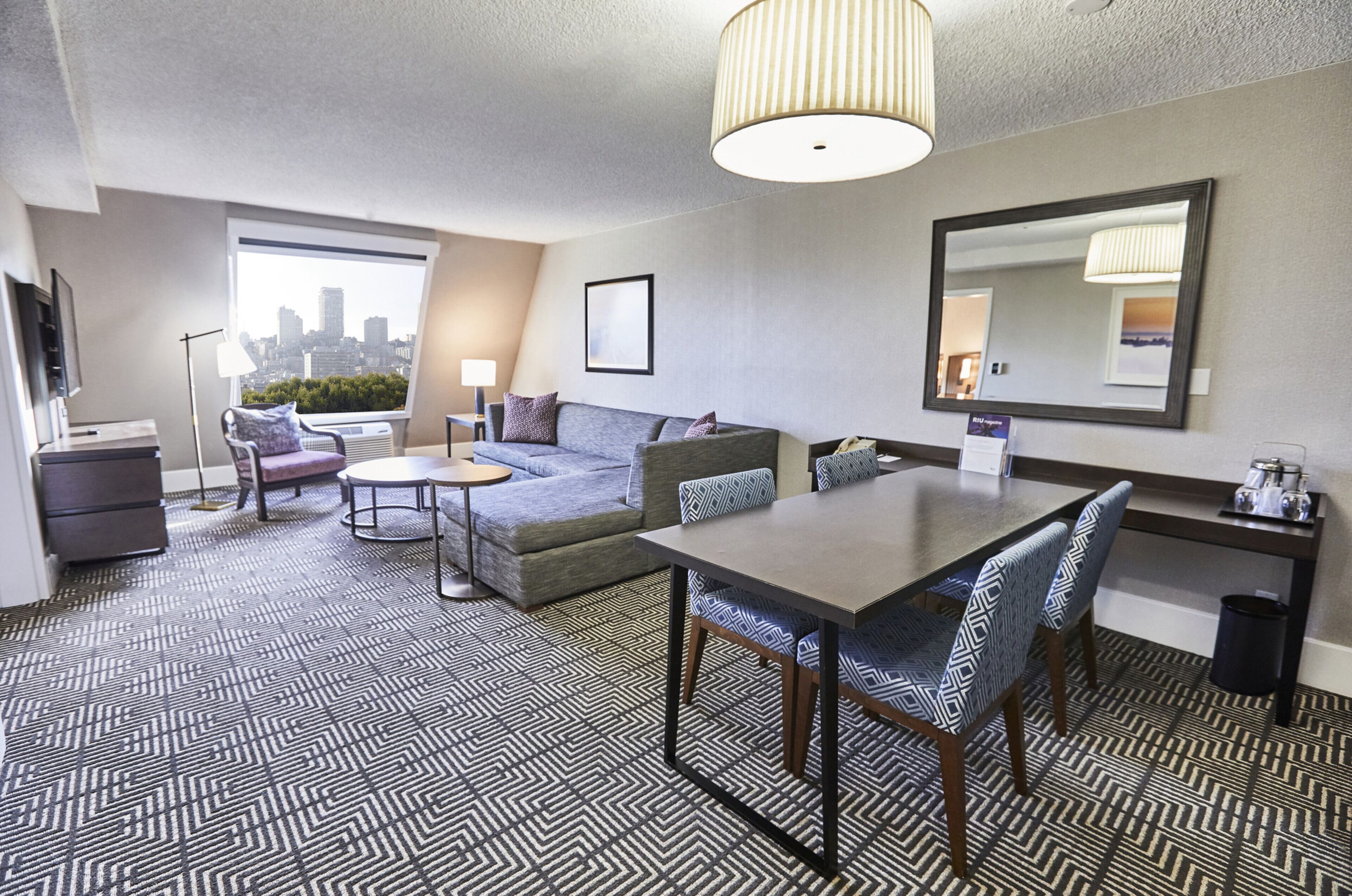 A hotel room with a sofa, a dining table, a TV, a lamp, and a large window with a city view.