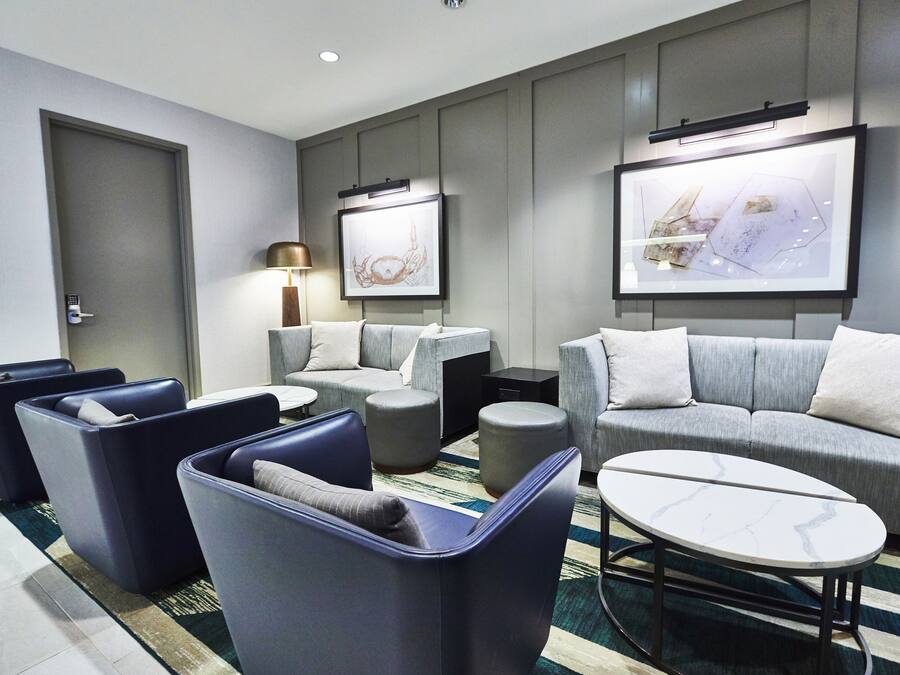 Ein modernes Hotelzimmer mit grauem Sofagarnitur, einem runden Couchtisch und einem großen, an der Wand befestigten Fernseher.