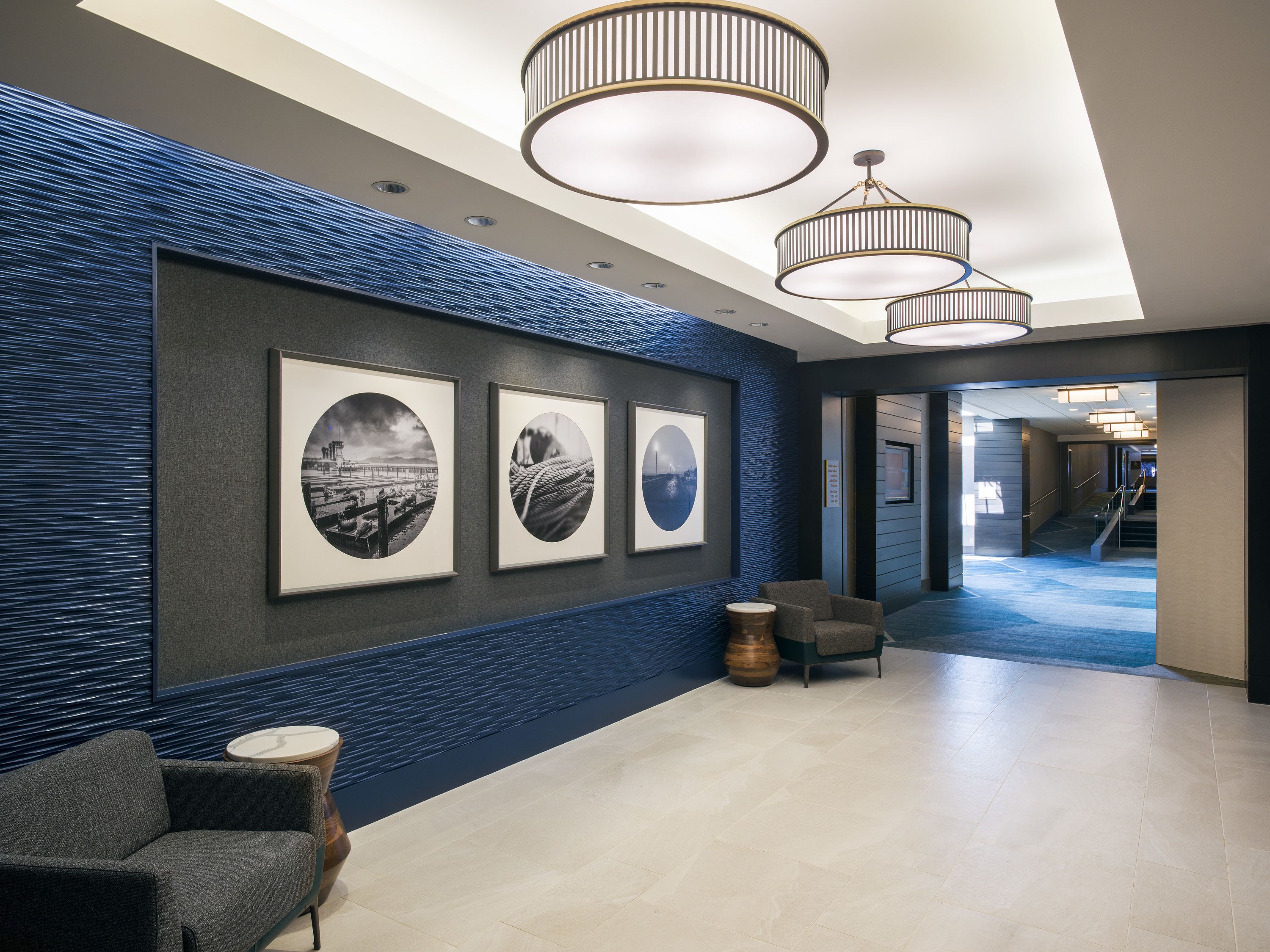 A modern lobby with a blue textured wall, framed black and white photographs, a grey sofa, and a round wooden table.