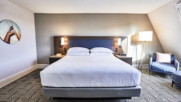 Una habitación de hotel con una cama grande, cabecera, mesitas de noche con lámparas, una silla y una lámpara de pie.