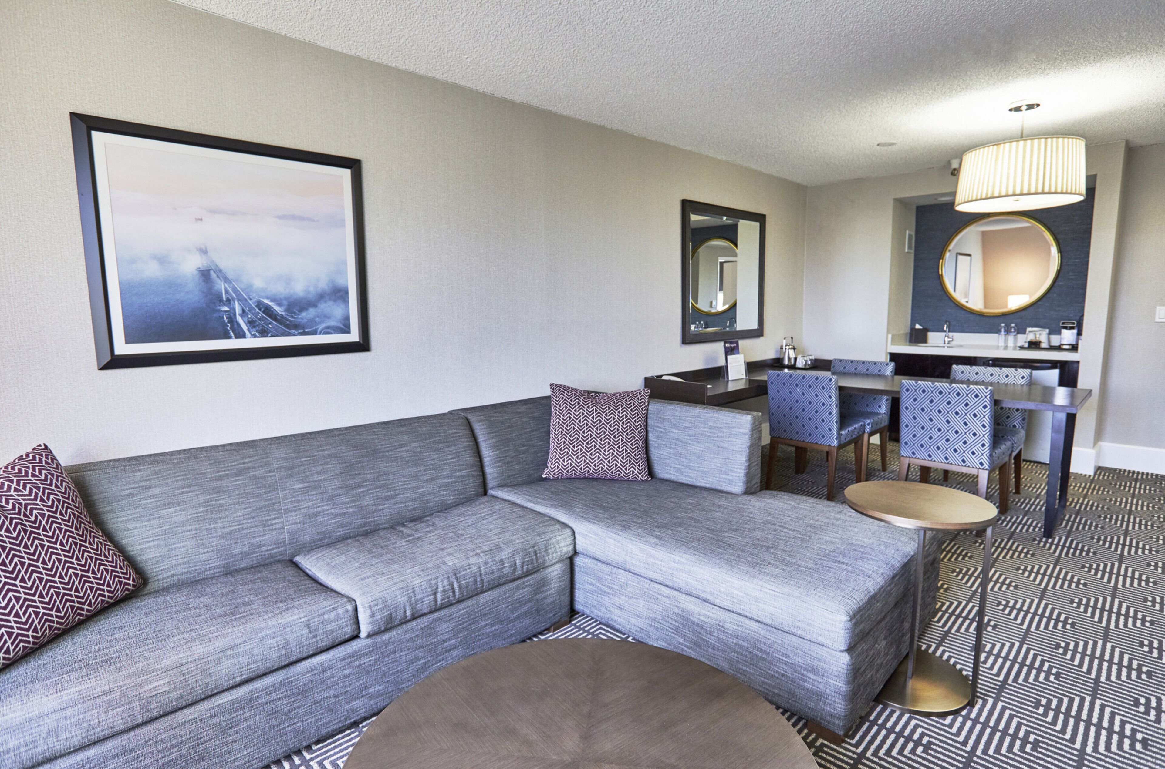 Una habitación de hotel moderna con un sofá seccional gris, una mesa de centro redonda, una zona de comedor con sillas azules y un espejo grande sobre el lavabo.