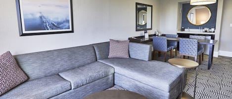 Una habitación de hotel moderna con un sofá seccional gris, una mesa de centro redonda, una zona de comedor con sillas azules y un espejo grande sobre el lavabo.