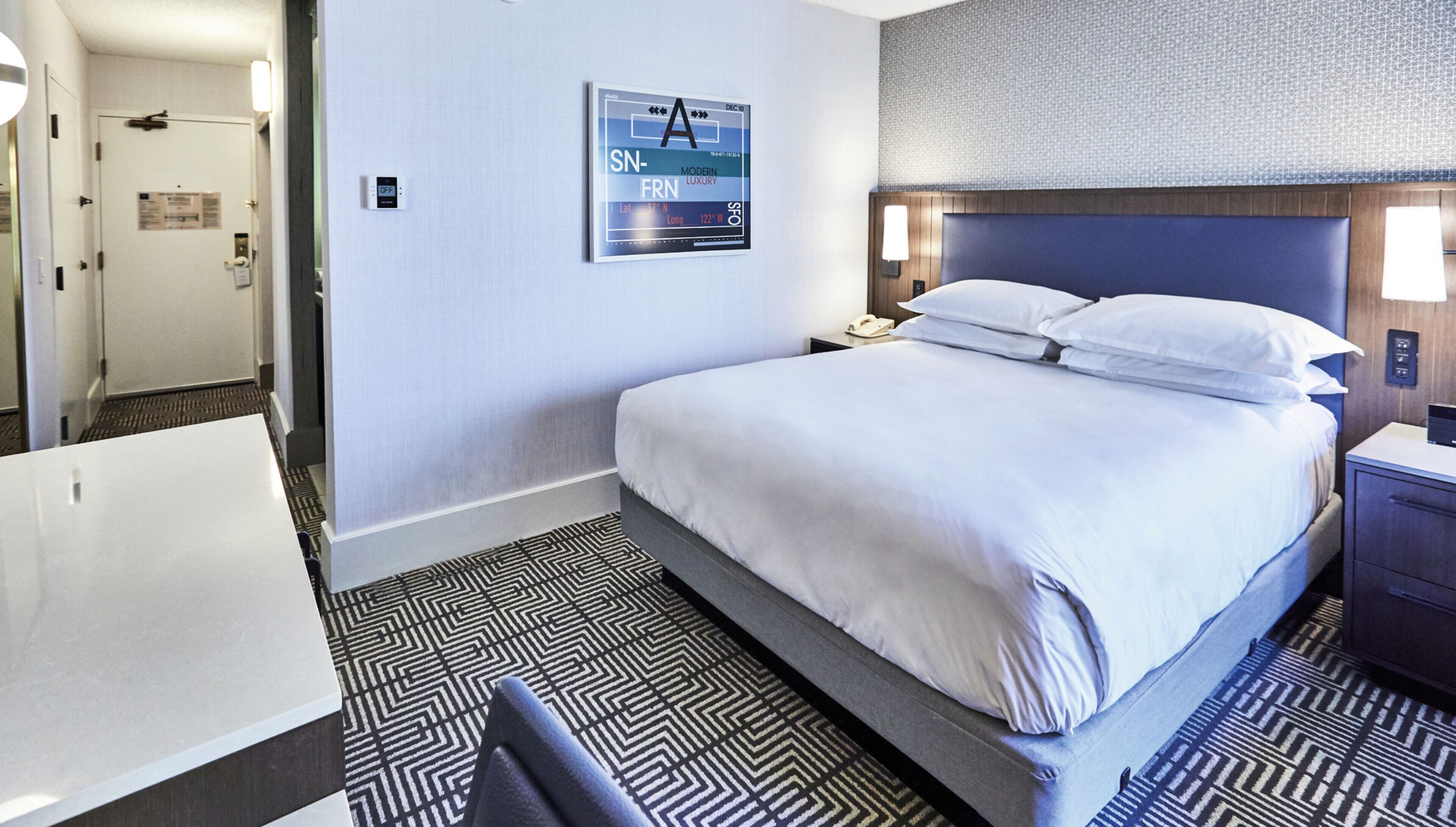 A hotel room with a bed, a nightstand, a lamp, a wall clock, and a poster on the wall.