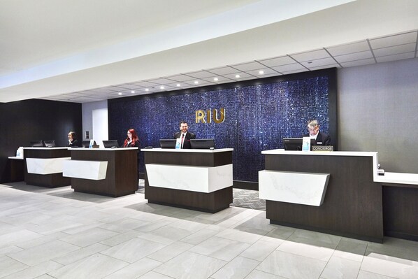 A modern reception area with multiple counters, a dark wall with 'RIU' branding, and a tiled floor.
