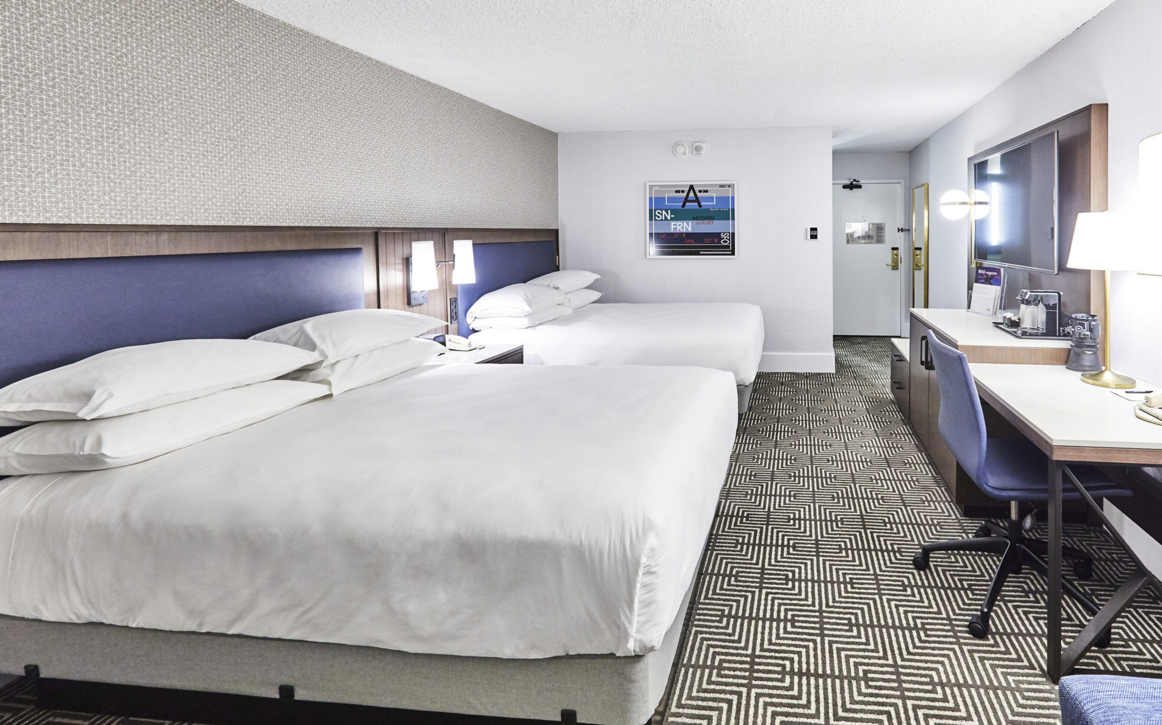 Habitación de hotel con dos camas, un escritorio, una silla, un espejo y un piso con diseño.