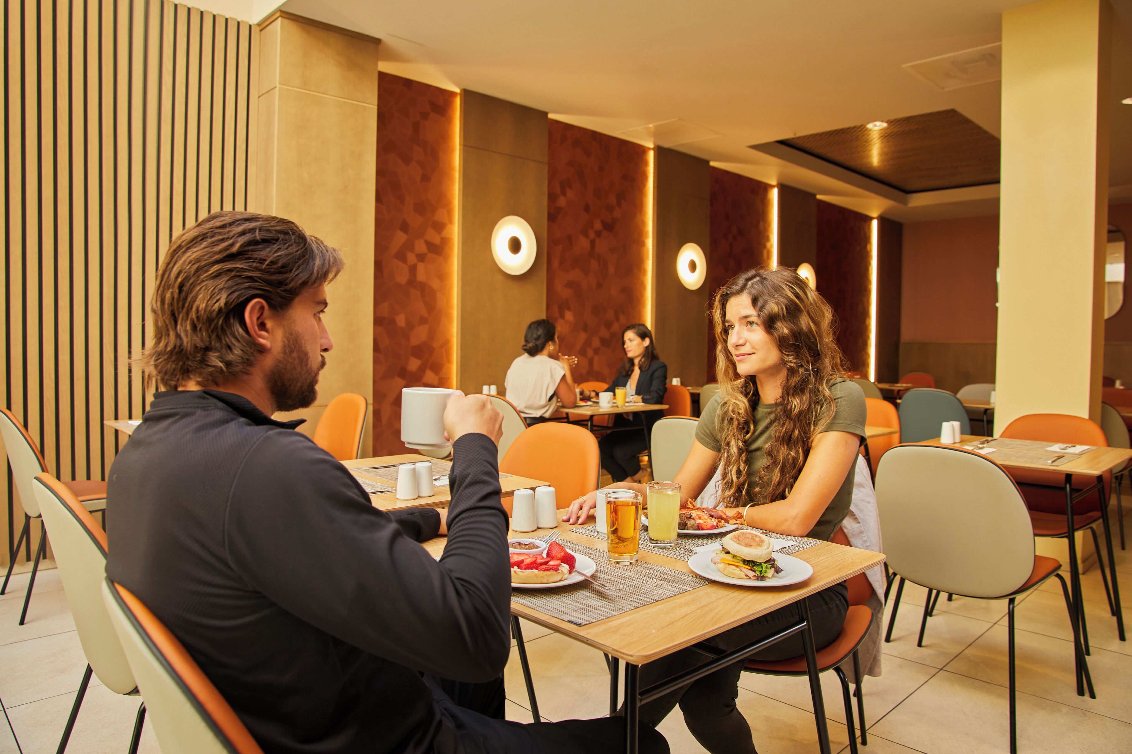 A dining area with tables and chairs, a person sitting at a table, and another person standing in the background.