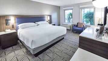 Habitación de hotel con una cama grande, un escritorio con silla, una mesita y una ventana con vistas a zonas verdes.