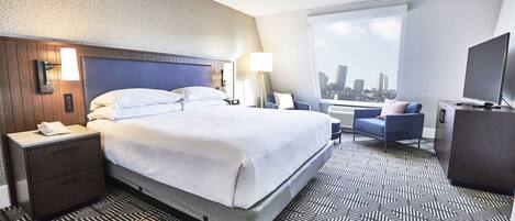 A hotel room with a large bed, a desk, a chair, a TV, and a window with a city view.