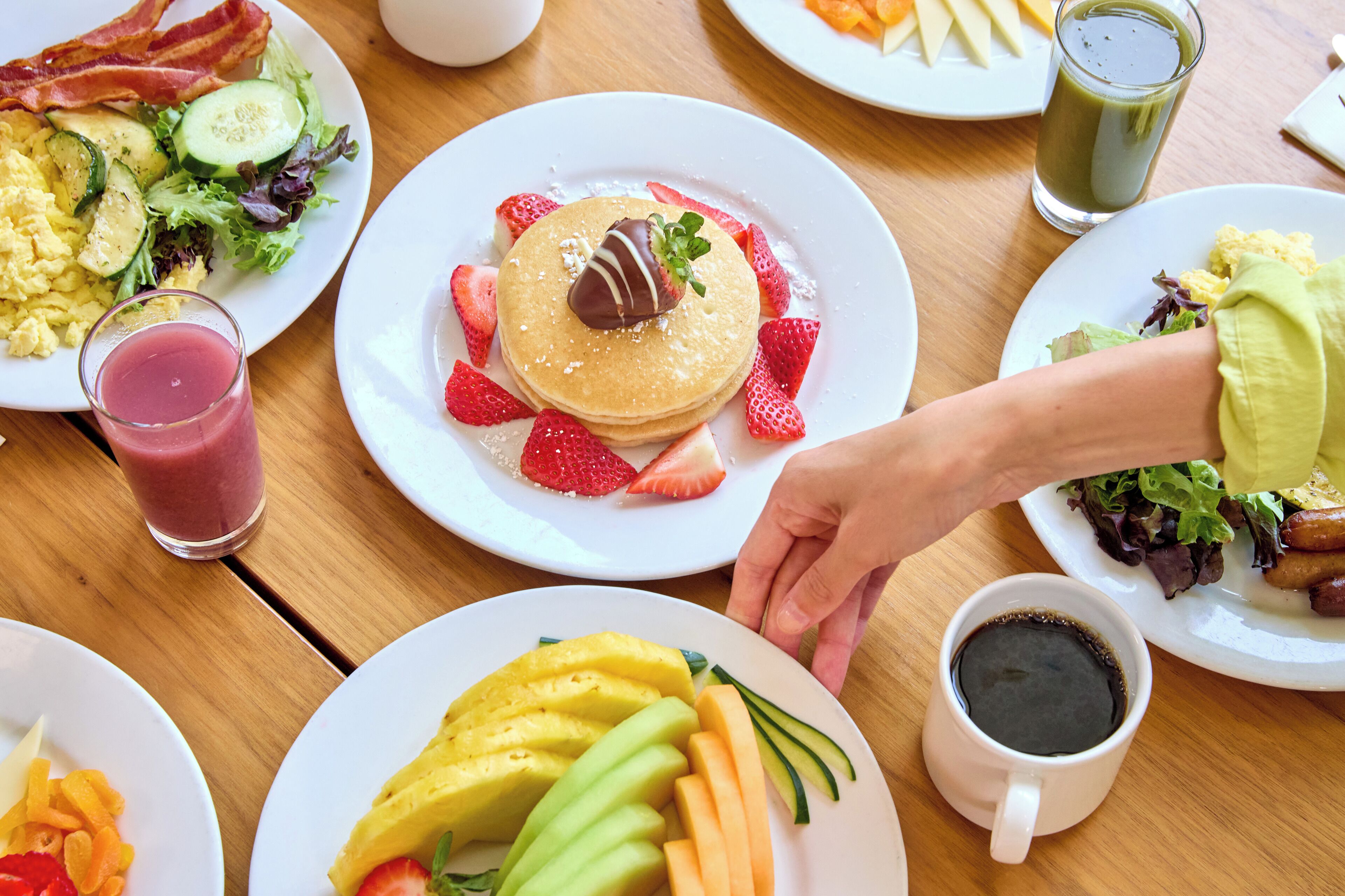 Een ontbijttafel met diverse gerechten, waaronder pannenkoeken, fruit, roerei, spek en een groene smoothie.