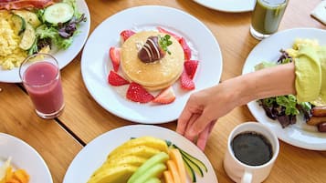 Una mesa de desayuno con diversos platos, entre ellos panqueques, fruta, huevos revueltos, tocino y un batido verde.