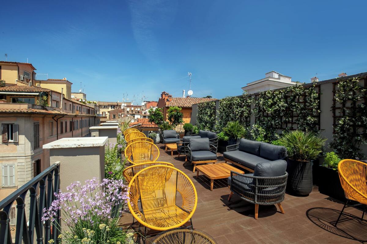 A rooftop terrace with outdoor seating, including wicker chairs and a sofa, surrounded by potted plants and a view of the cityscape.