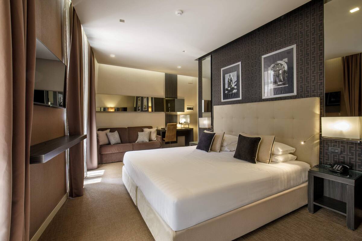 A modern hotel room with a large bed, a desk, a sofa, and a mirror.