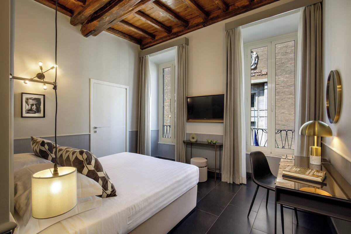 A bedroom with a bed, bedside lamp, TV, desk, chair, and a view of the outside.