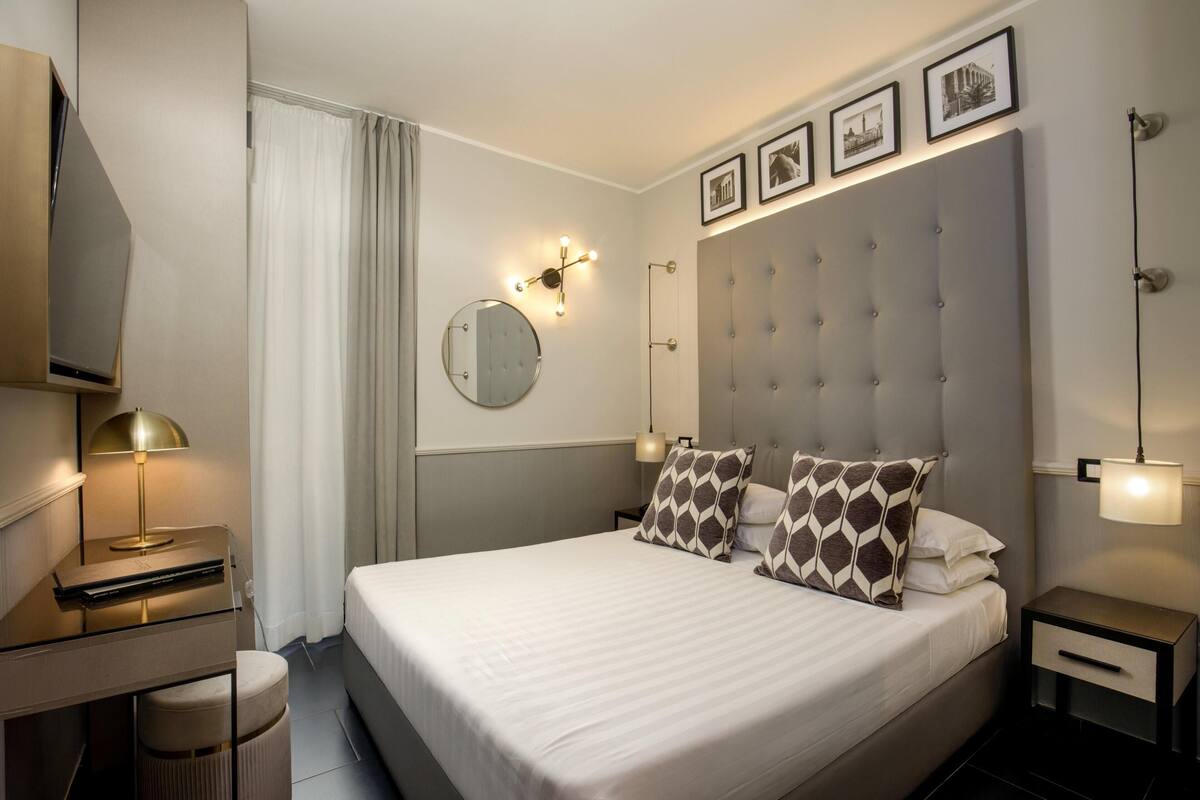 A modern hotel room with a large bed, a desk, a chair, a round mirror, and a nightstand with a lamp.