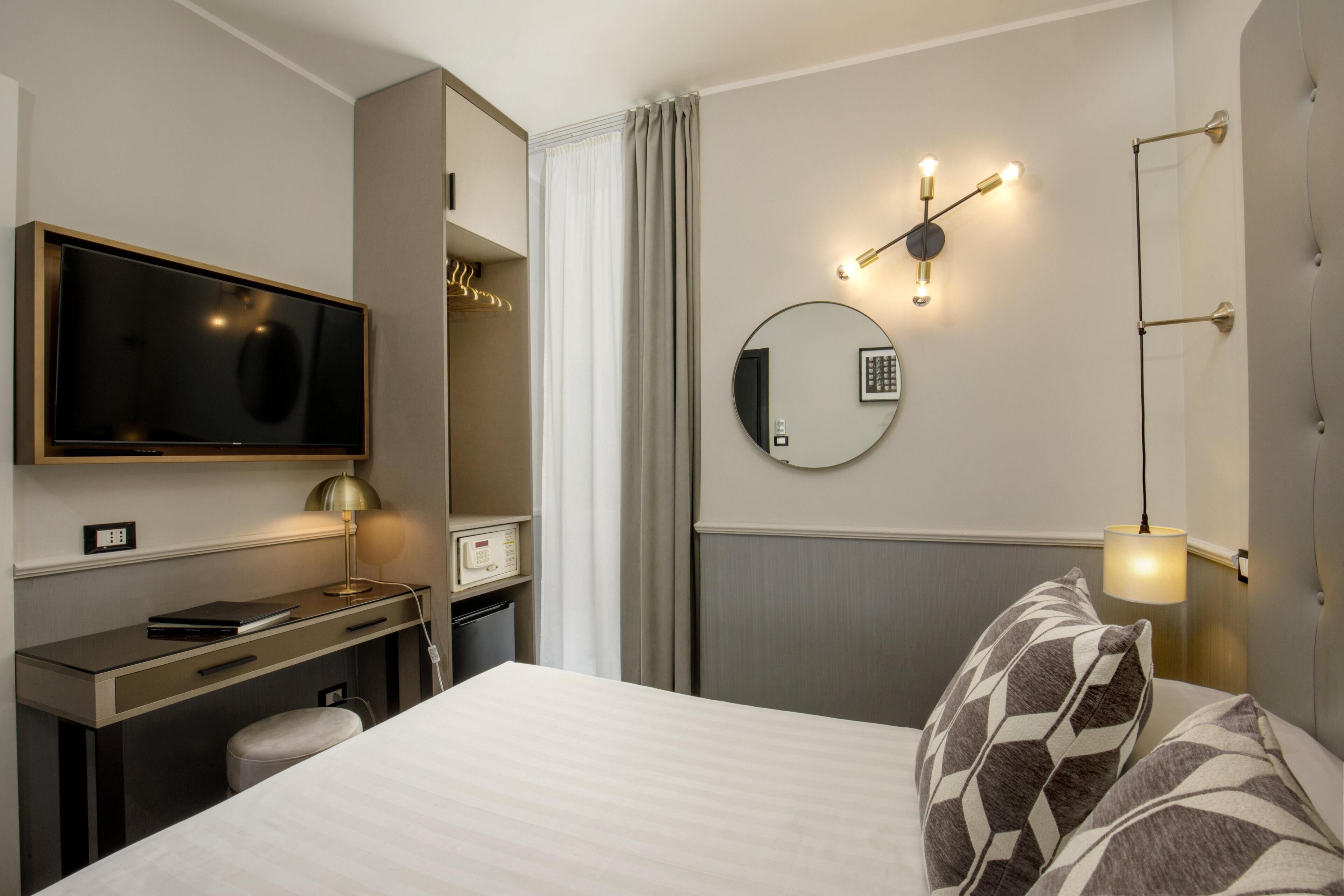 A hotel room with a bed, a desk with a computer, a television, a round mirror, and a wall-mounted light fixture.