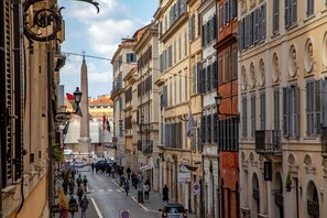 A street lined with historic buildings, a prominent obelisk, and pedestrians.