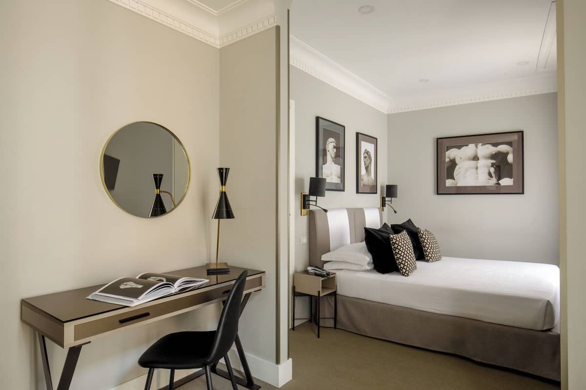 A hotel room with a bed, a desk with a chair, a round mirror, and framed artwork on the wall.