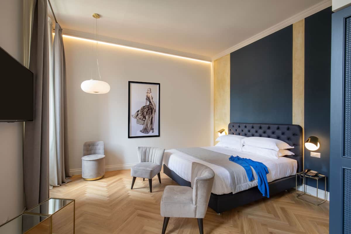 A modern bedroom with a large bed, two chairs, a side table, and a framed artwork on the wall.