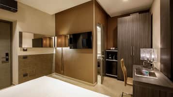 A modern hotel room with a large bed, a desk with a lamp, a chair, and a wardrobe.