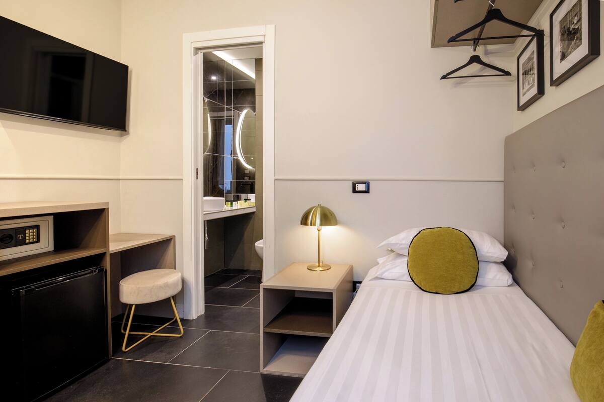 A hotel room with a bed, a desk, a chair, a TV, and a bathroom visible through an open door.
