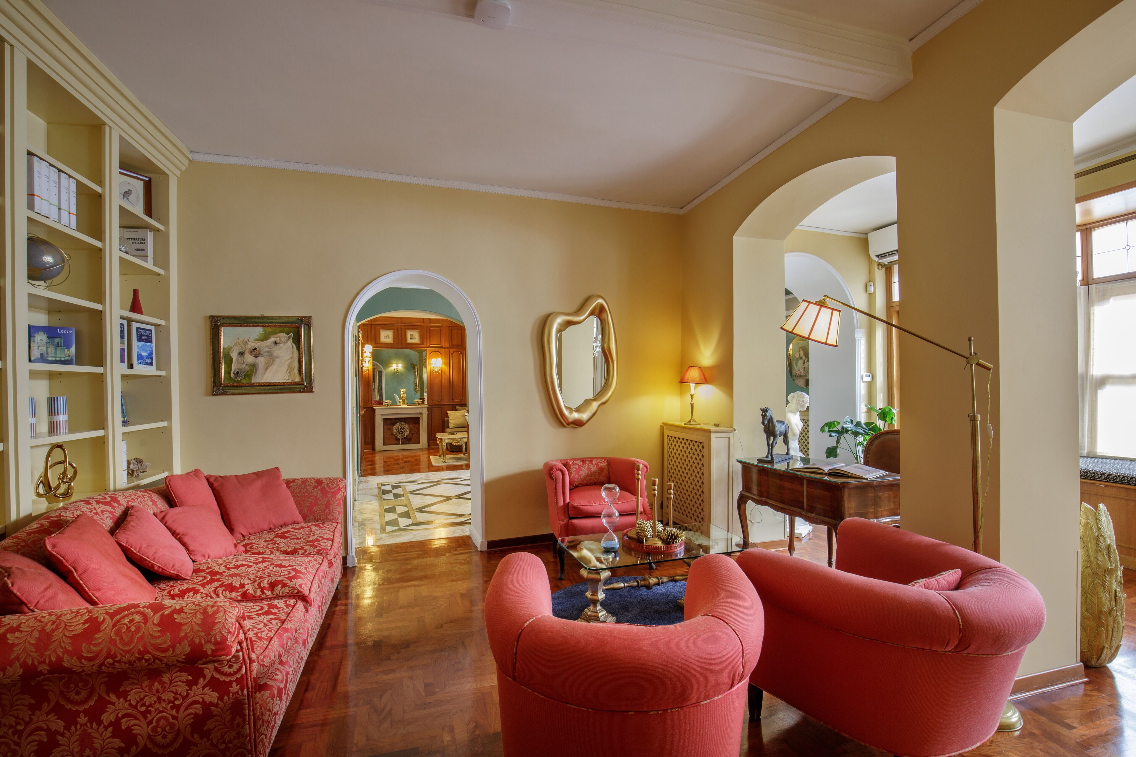 A spacious living room with a red sofa, two red armchairs, a bookshelf, and a decorative mirror.