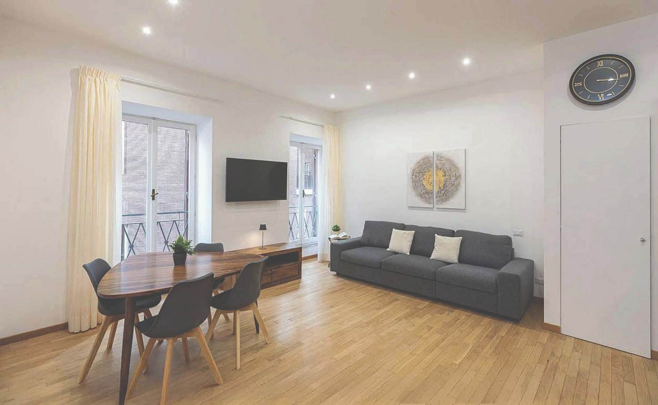A modern living room with a dining area, a grey sofa, a flat-screen TV, and a clock on the wall.