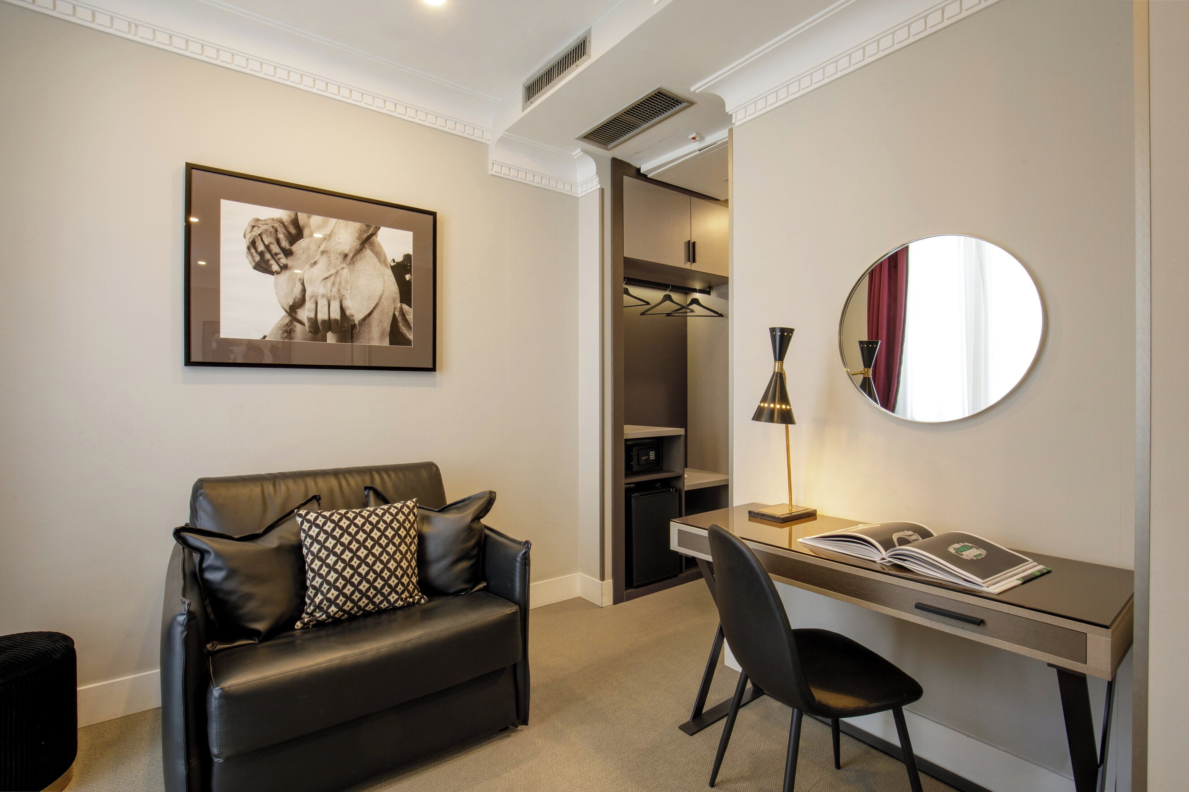 A modern hotel room with a leather sofa, a desk with a chair, a framed artwork, a mirror, and a wardrobe.