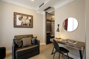 A modern hotel room with a leather sofa, a desk with a chair, a framed artwork, a mirror, and a wardrobe.