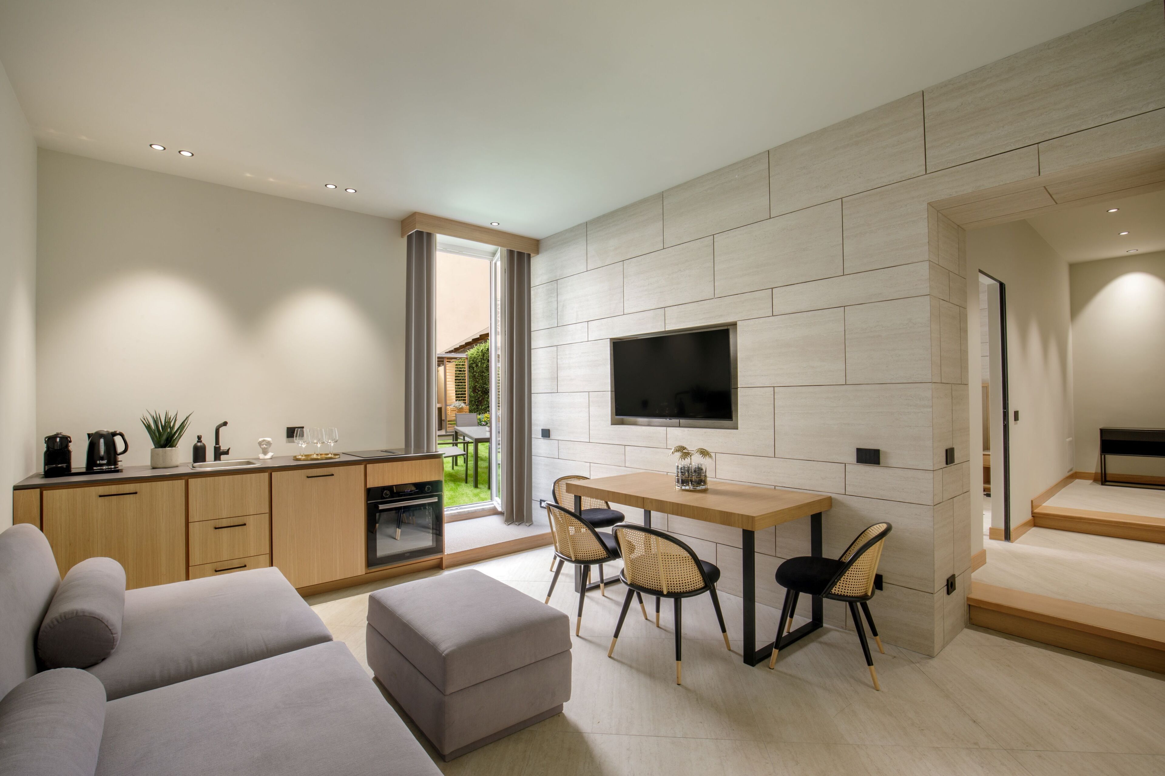 A modern living room with a sofa, a dining table with chairs, a kitchen area with cabinets, and a TV mounted on the wall.