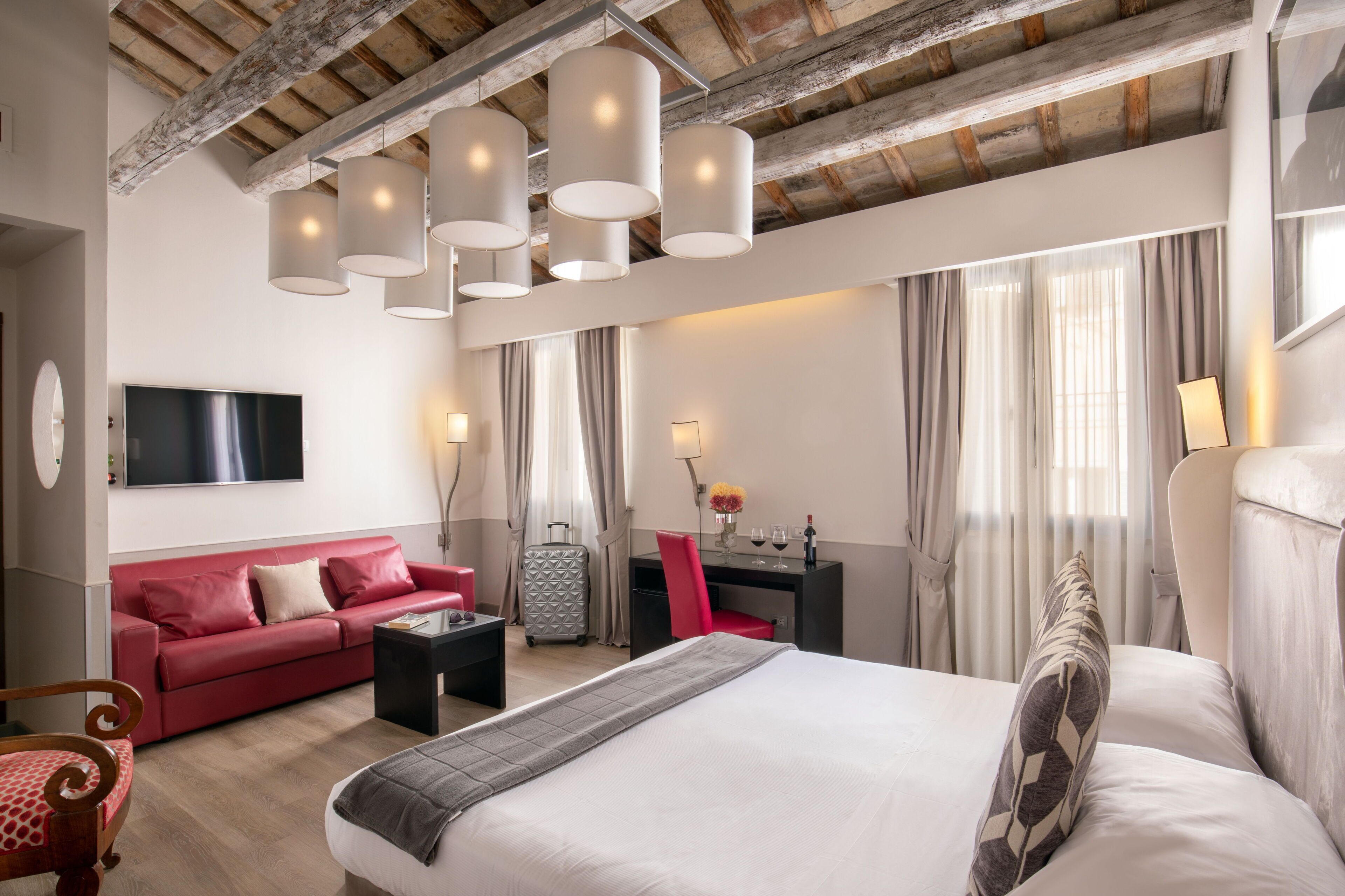 A hotel room with a large bed, a red sofa, a desk with a chair, a TV mounted on the wall, and a ceiling with exposed beams.