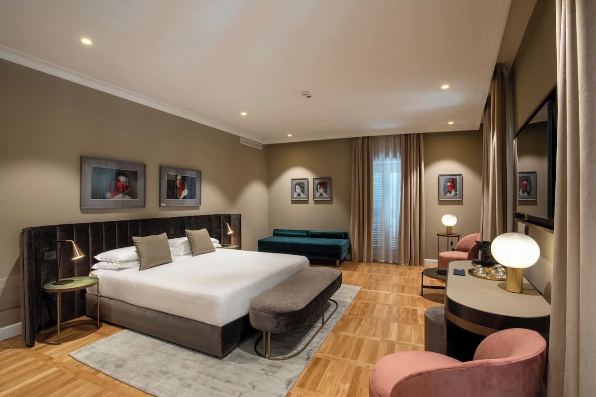 A modern hotel room with a large bed, a seating area with a sofa and armchairs, a small table, and a desk with a lamp.