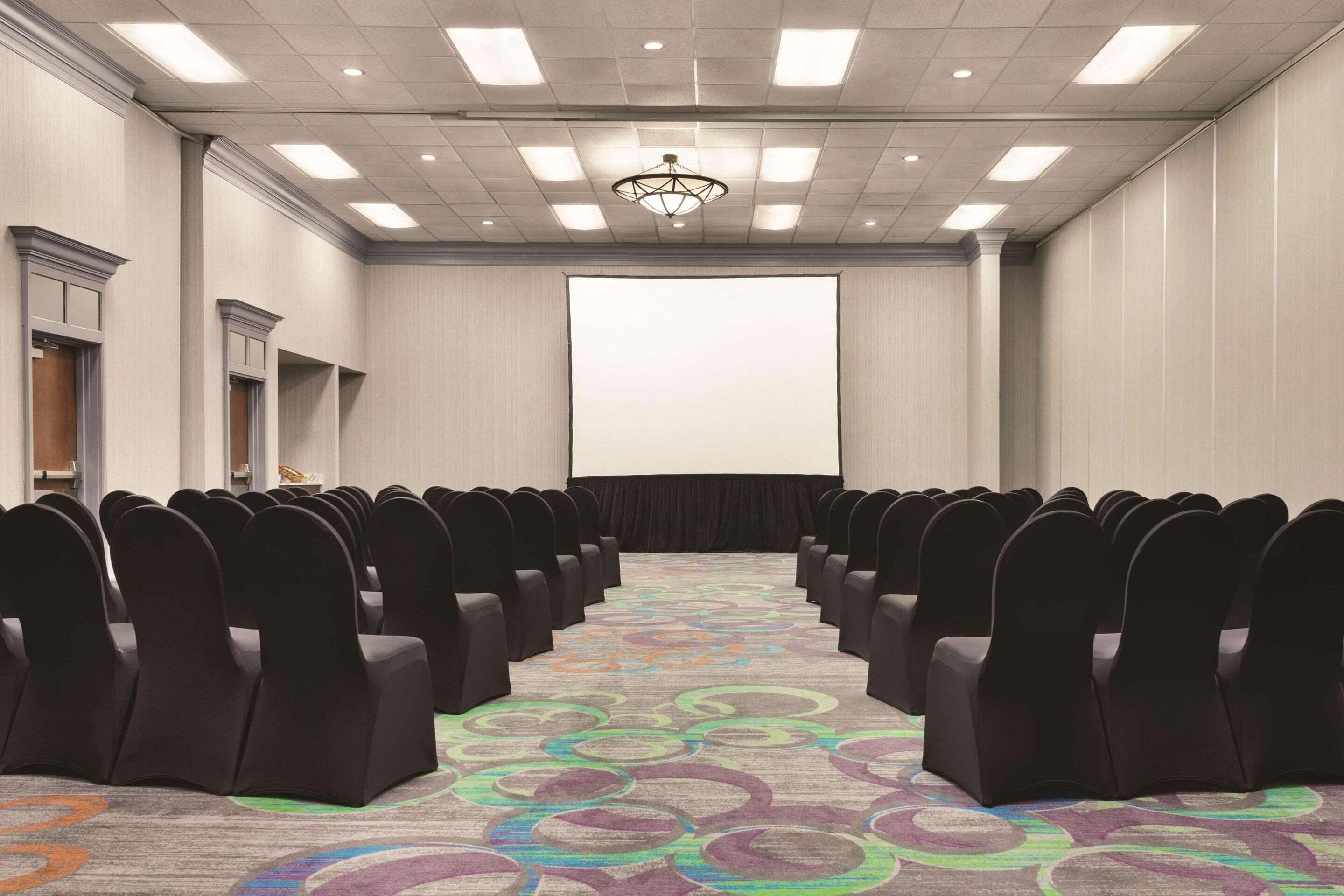 Un salón de conferencias con filas de sillas, una pantalla de proyector y una alfombra con diseños circulares.