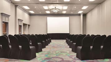 Un salón de conferencias con filas de sillas, una pantalla de proyector y una alfombra con diseños circulares.