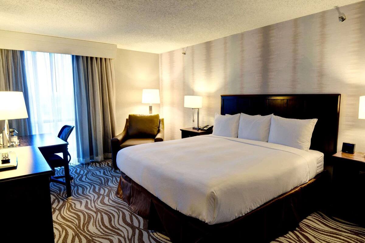 A hotel room with a large bed, two bedside tables with lamps, a desk with a chair, and a large window with curtains.