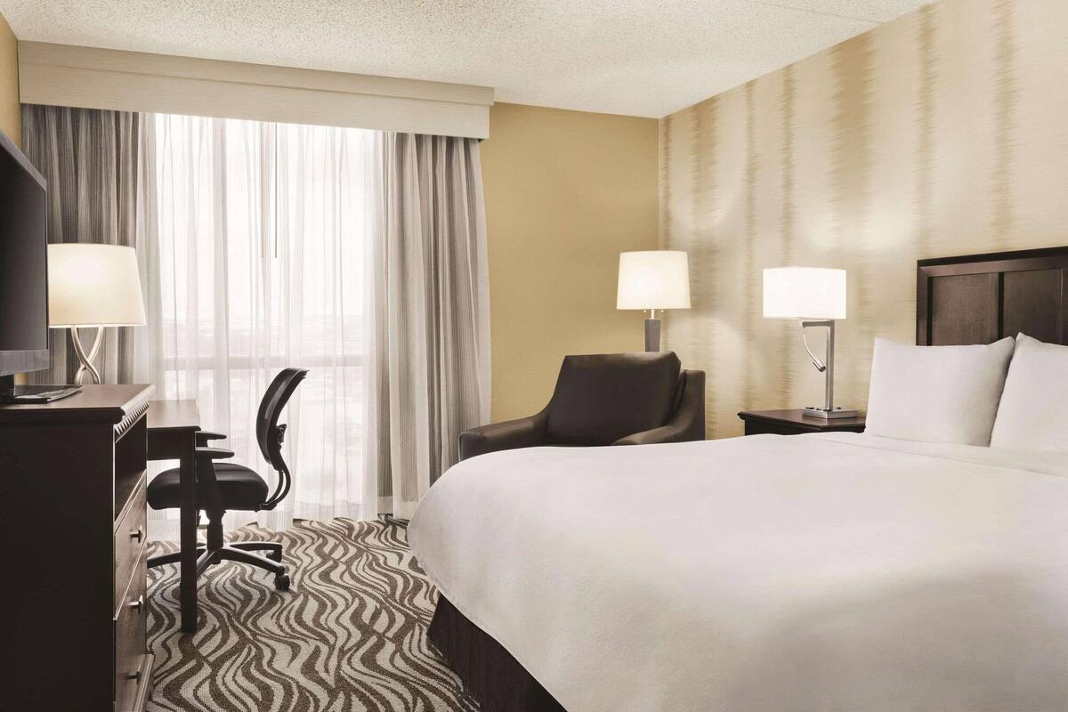 A hotel room with a large bed, two bedside tables with lamps, a desk with a chair, and a view through sheer curtains.