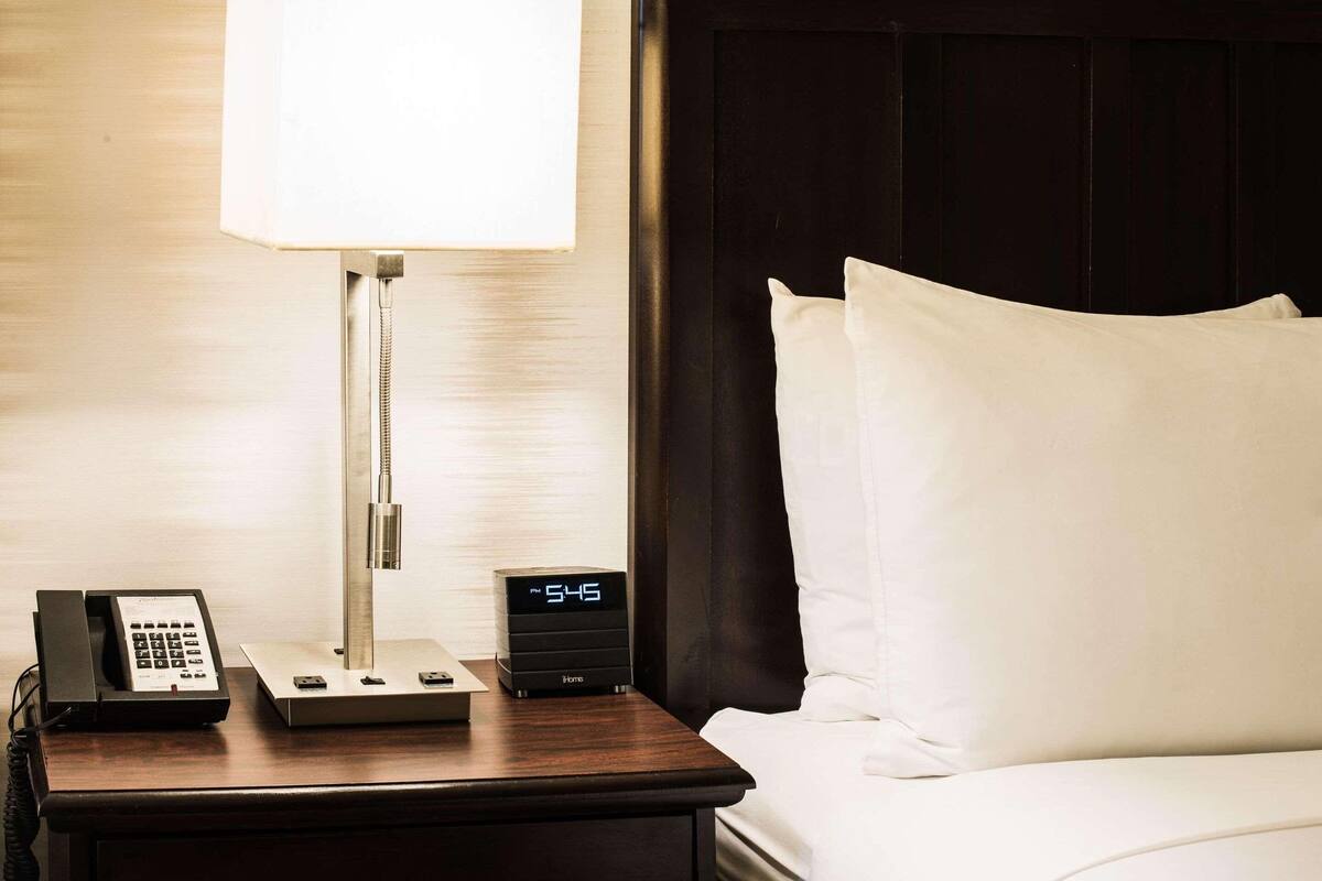 A hotel room with a bedside table featuring a lamp, a digital clock, and a telephone.