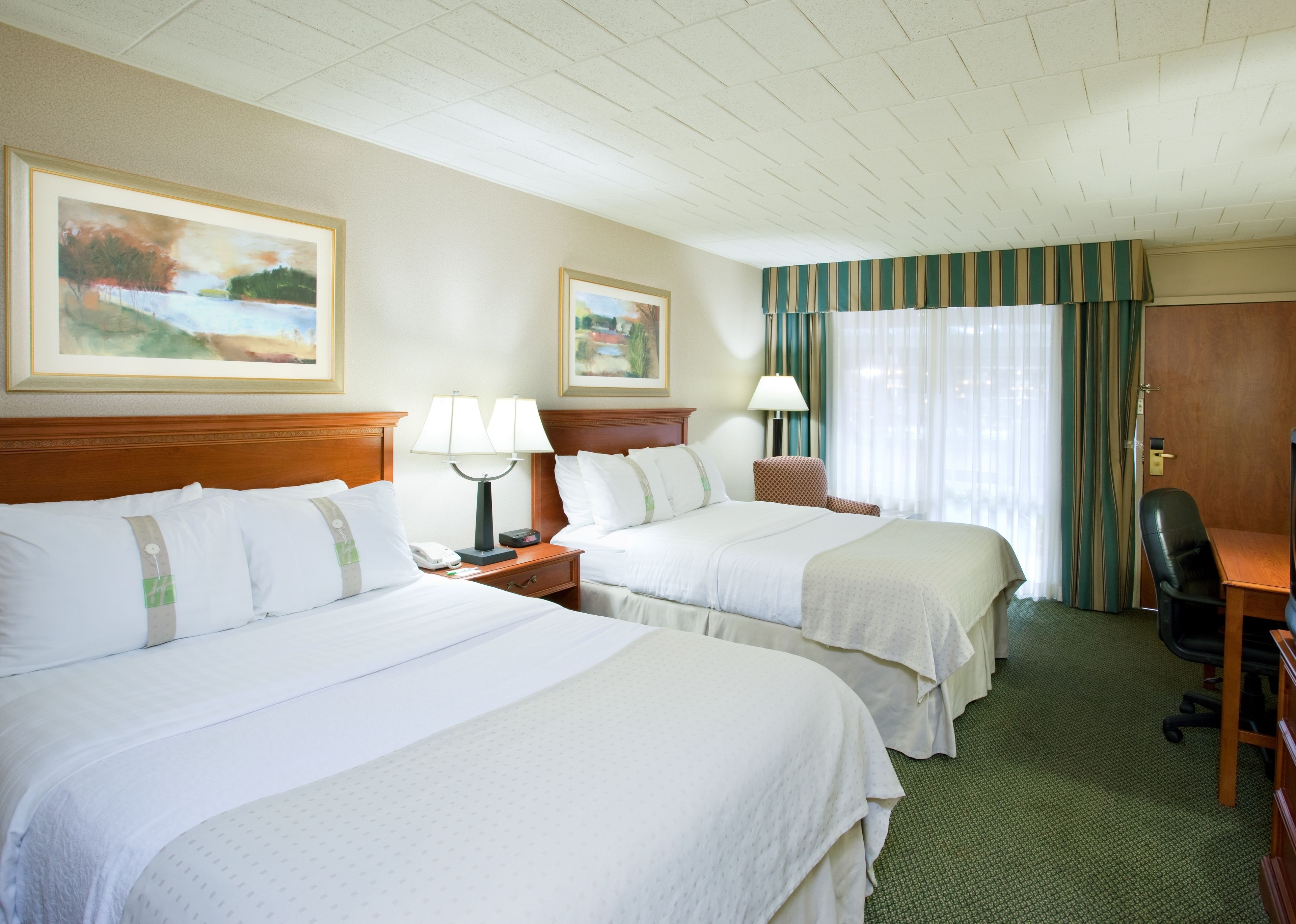A hotel room with two beds, a desk, a chair, a lamp, and two paintings on the wall.
