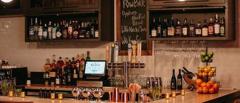 Un espace bar avec un tableau noir affichant le menu, où figurent des articles tels que des huîtres et des « blue point ». On y trouve des étagères remplies de bouteilles et de verres, une caisse enregistreuse et un présentoir de fruits.
