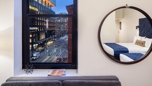 A modern hotel room with a large window offering a city view, a round mirror, and a neatly made bed.
