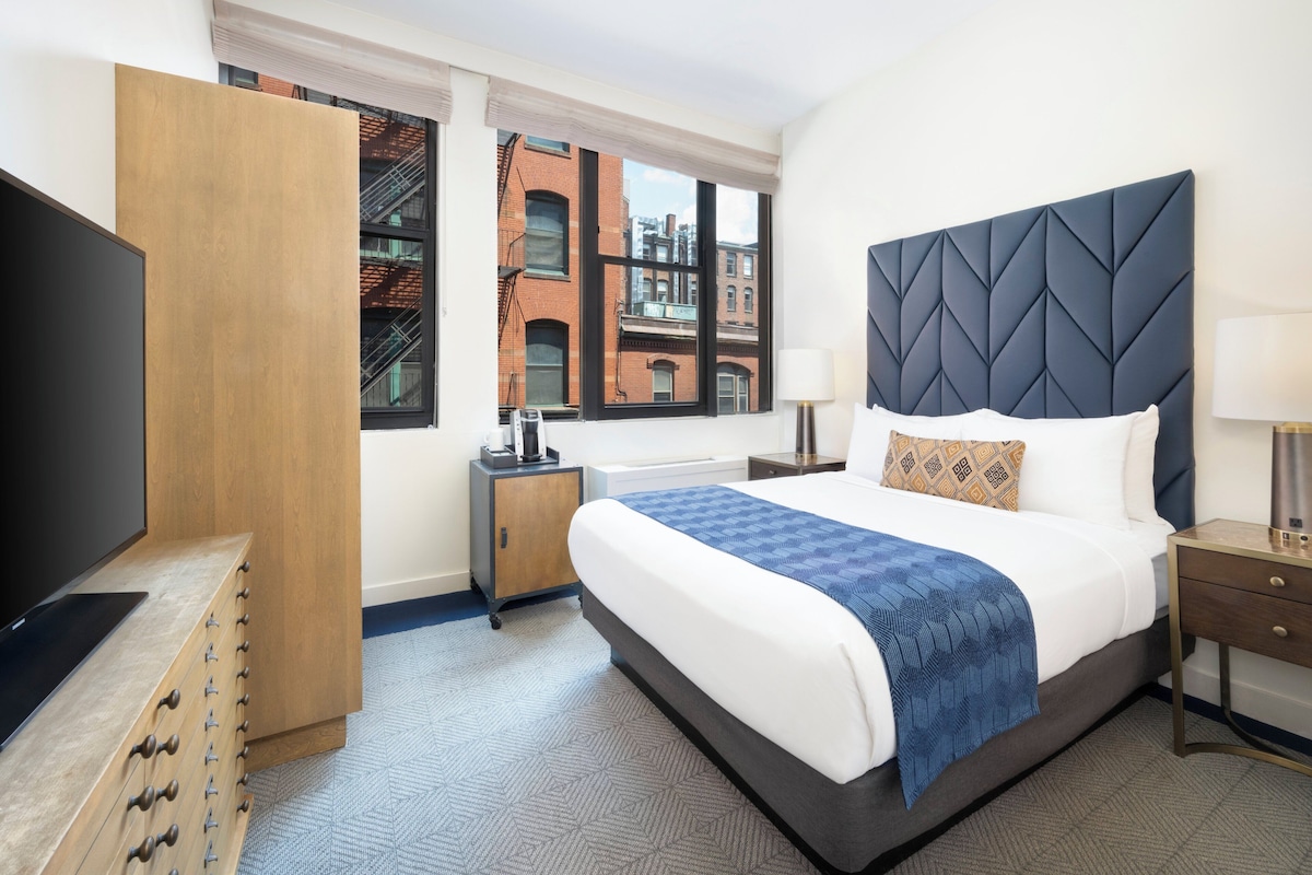 A hotel room with a large bed, a television, a wooden dresser, a nightstand, and a window with a city view.