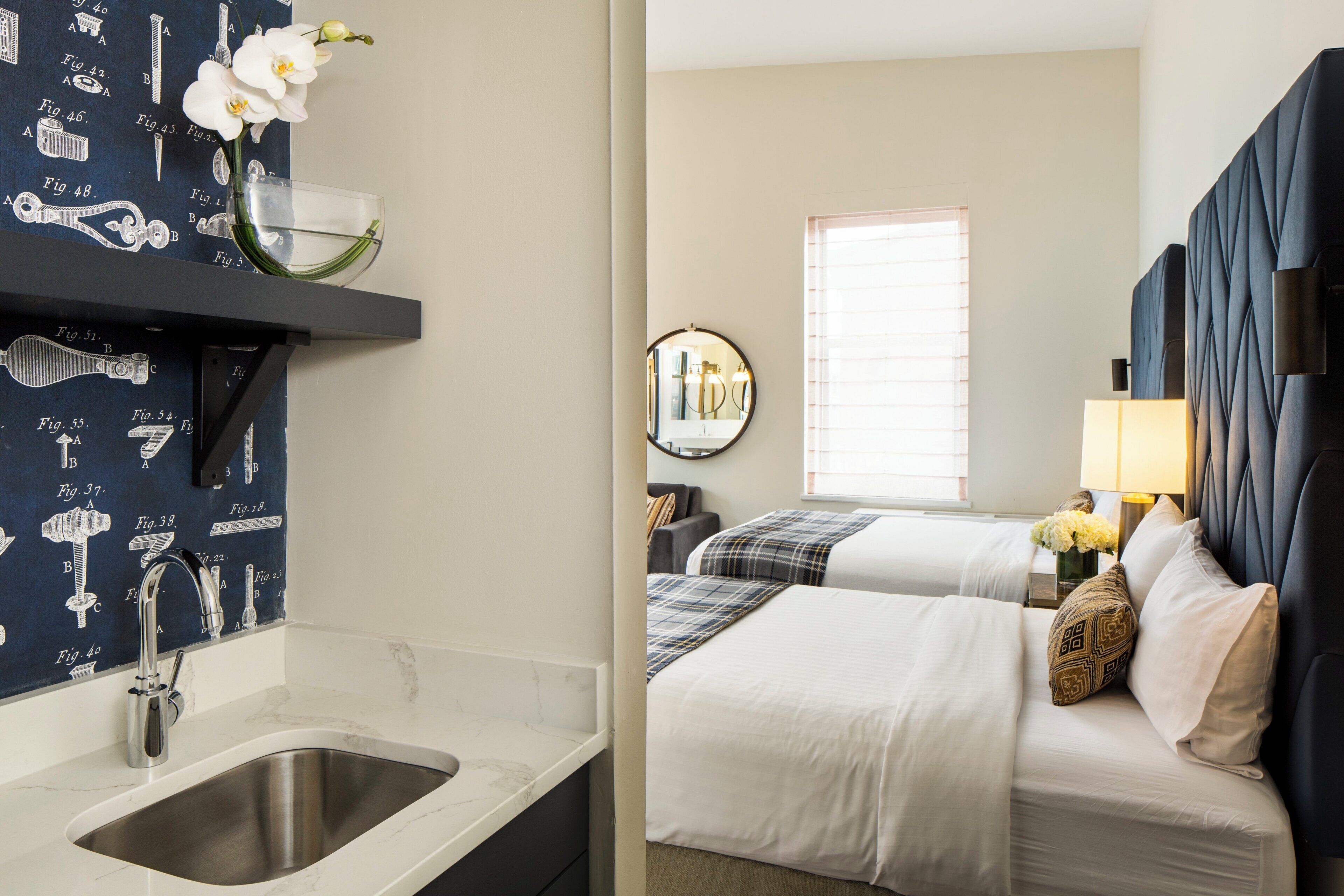A hotel room with a bathroom, a bed, a bedside table with a lamp, and a window with blinds.