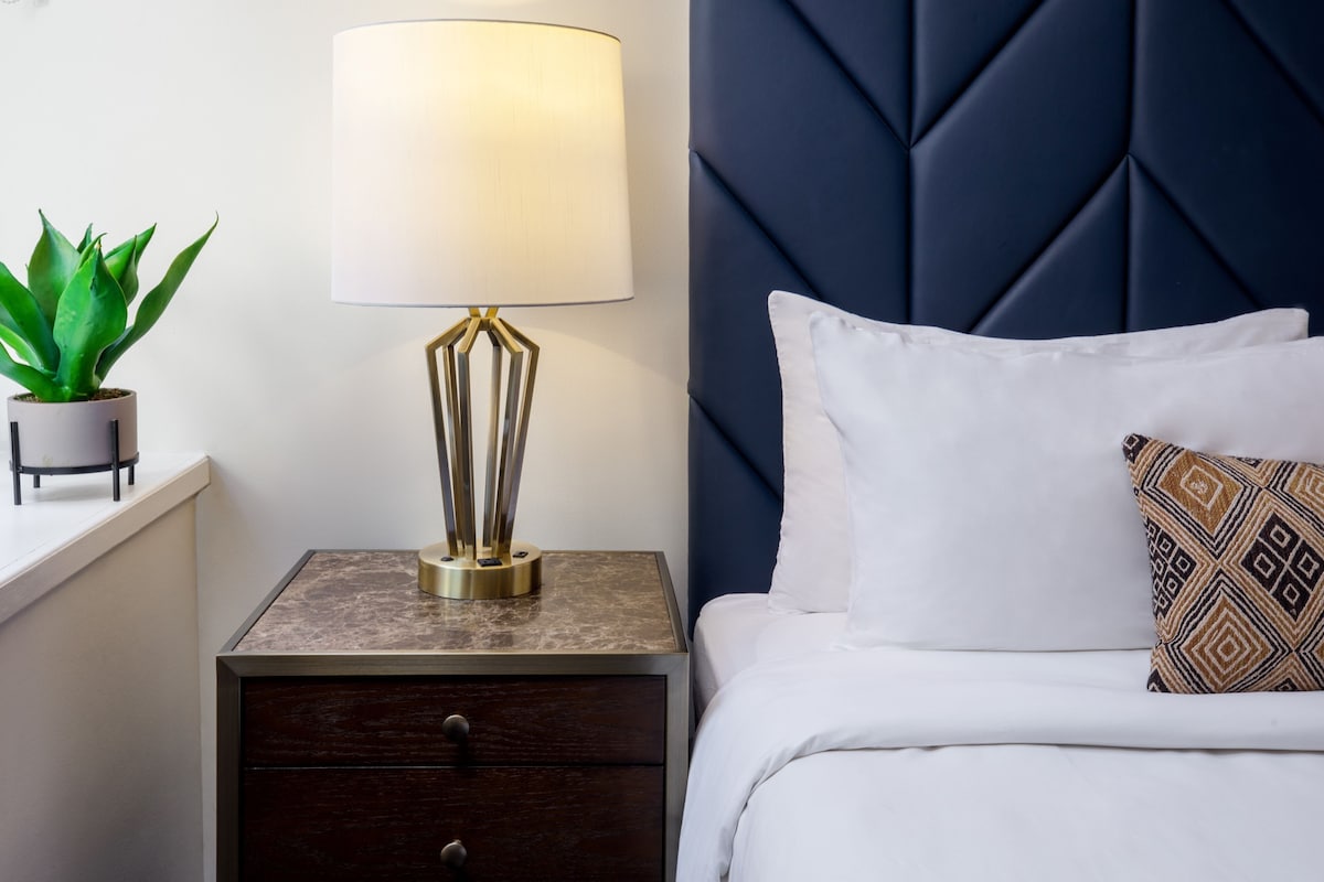A neatly made bed with white bedding and a patterned pillow, a bedside lamp, a small potted plant, and a dark wooden nightstand with a drawer.