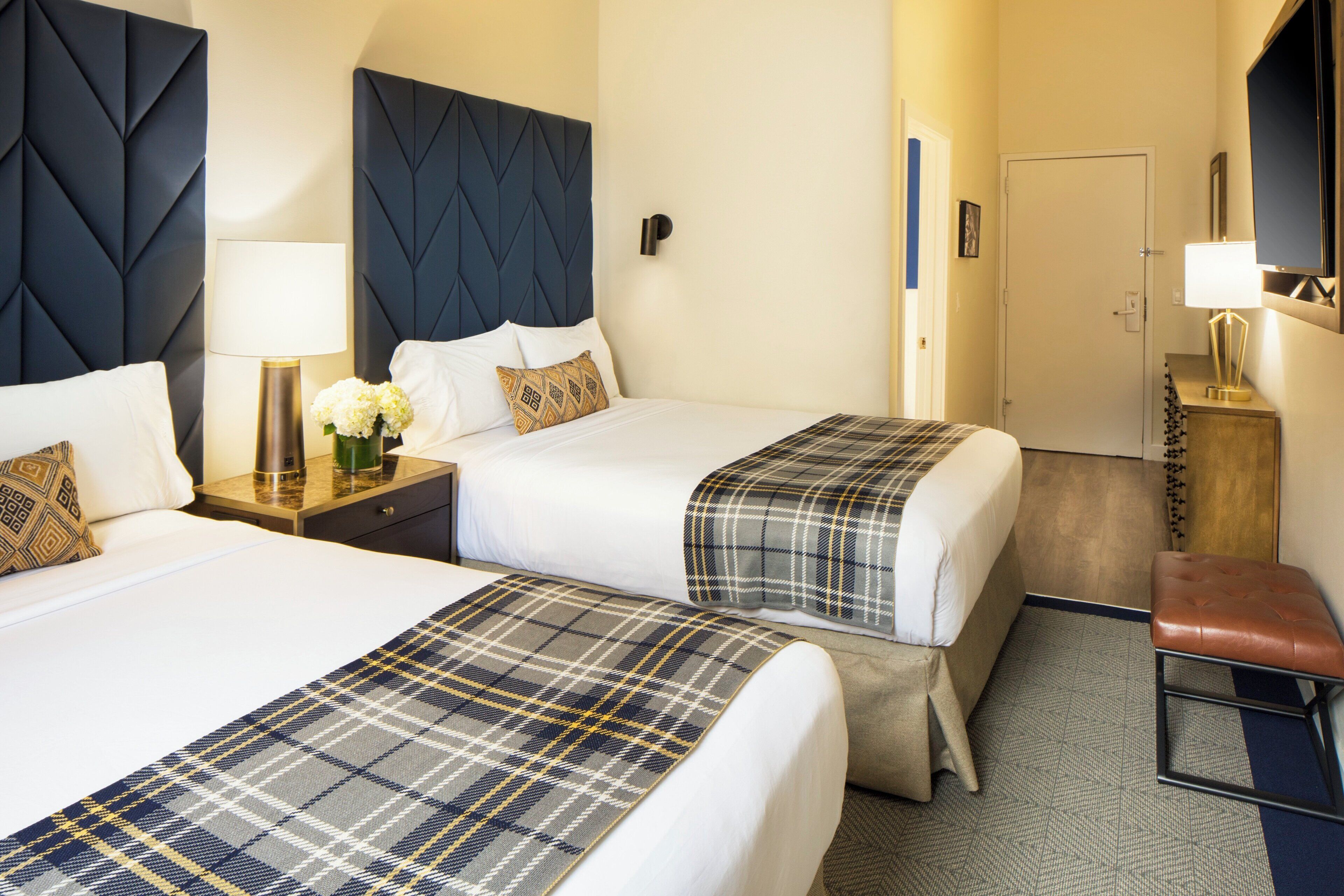 A hotel room with two beds, a nightstand with a lamp, a small table with a vase of flowers, and a television on the wall.