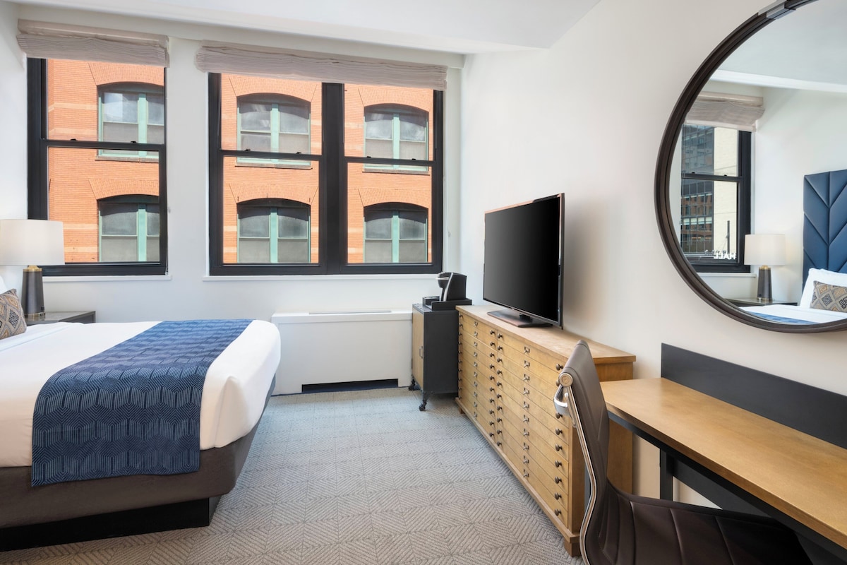 A hotel room with a bed, a desk, a television, a dresser, and a large window with a view of a brick building.