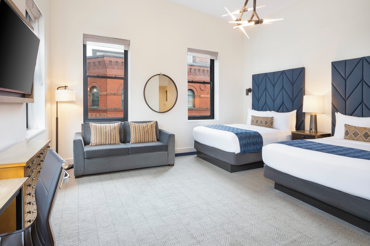 A hotel room with two beds, a sofa, a TV, and a large window with a view of a brick building.