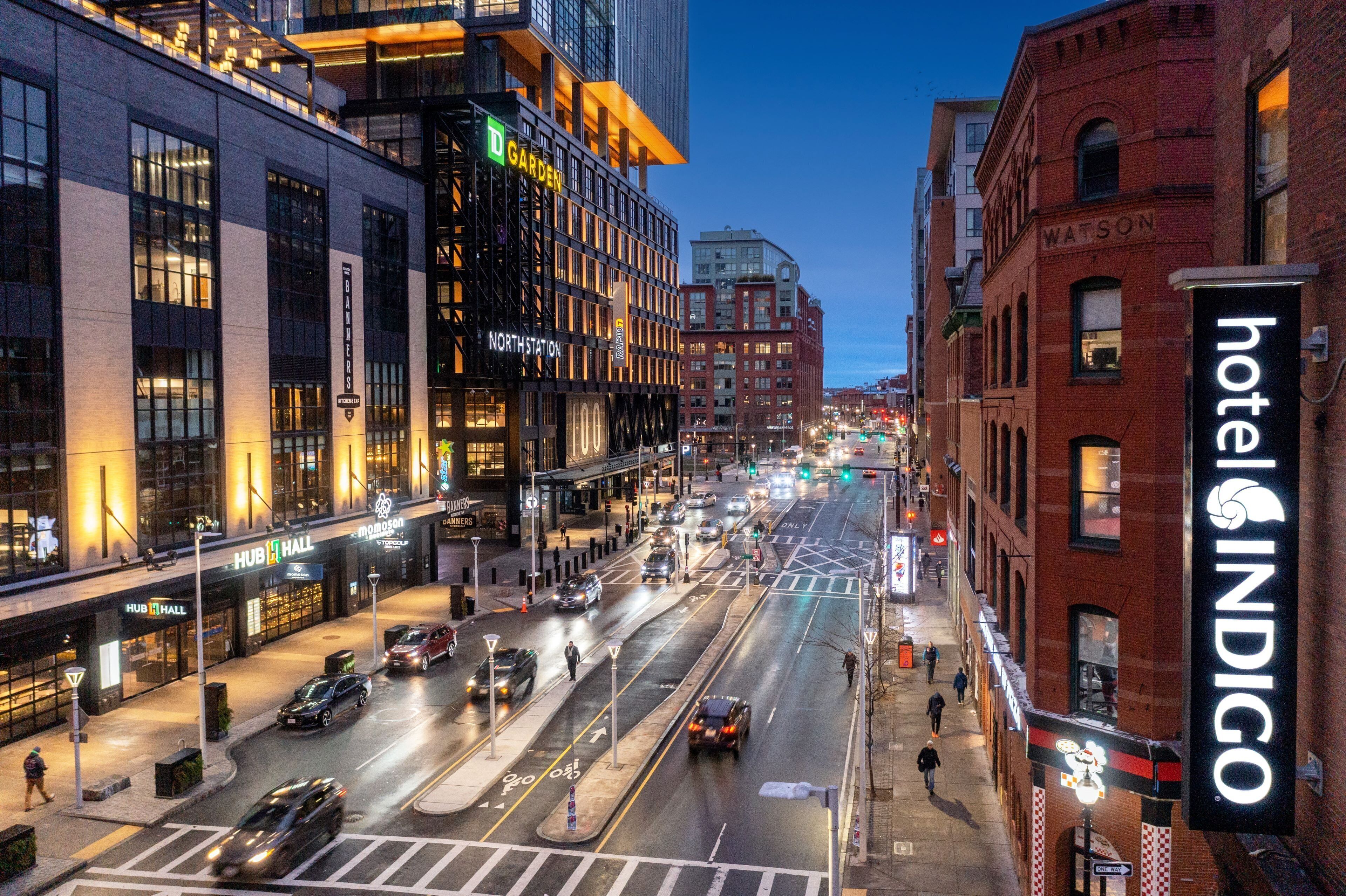 A city street at dusk with buildings, a hotel named 'hotel INDIGO', and a 'NORTHSTATION' sign.