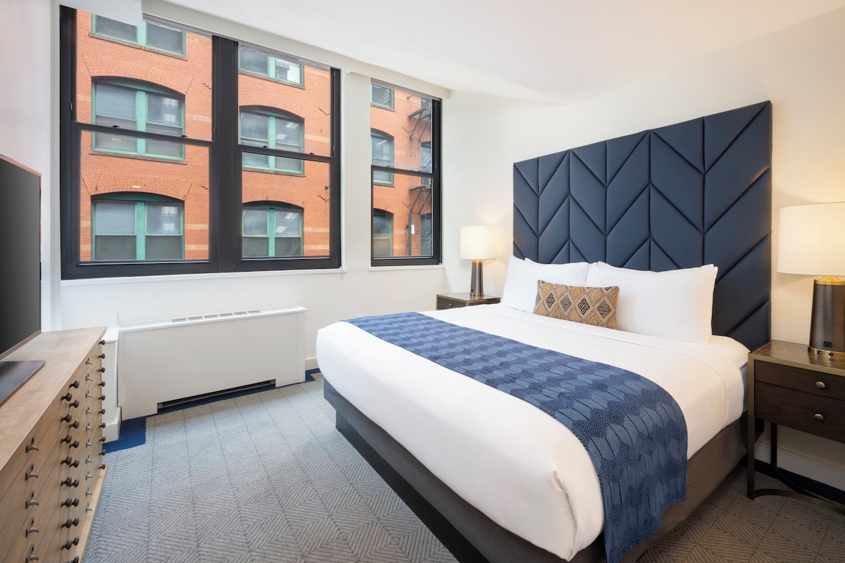 A hotel room with a large bed, a modern headboard, a dresser, and a window with a view of a brick building.