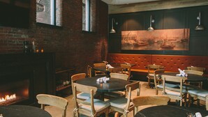 A restaurant with brick walls, wooden tables, and chairs. There is a fireplace with a fire, and a large painting on the wall.